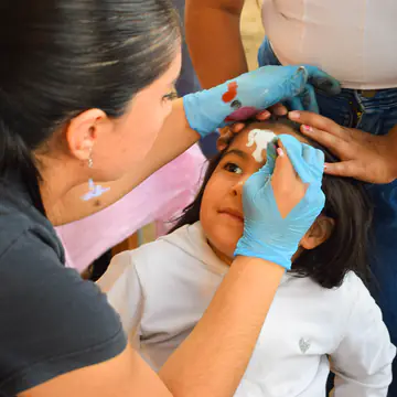 Manos con guantes pintan la frente de una nena que espera sentada.